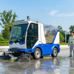Un véhicule de nettoyage urbain bleu et blanc avec balais, à côté de deux agents en uniformes de travail souriants, devant des immeubles modernes.