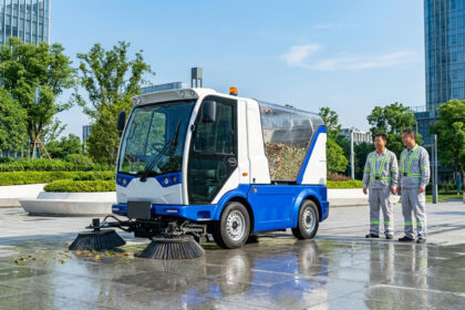 Un véhicule de nettoyage urbain bleu et blanc avec balais, à côté de deux agents en uniformes de travail souriants, devant des immeubles modernes.