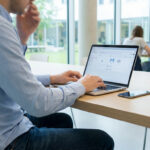 Homme travaillant sur un ordinateur portable affichant une interface d'e-mail et calendrier dans un espace lumineux. Un smartphone à côté.