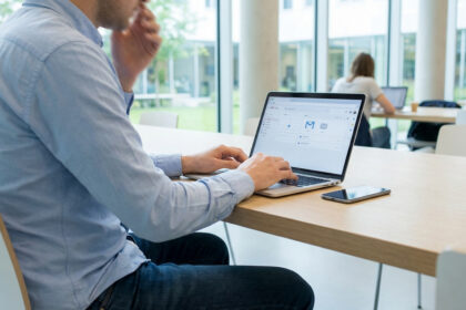 Homme travaillant sur un ordinateur portable affichant une interface d'e-mail et calendrier dans un espace lumineux. Un smartphone à côté.